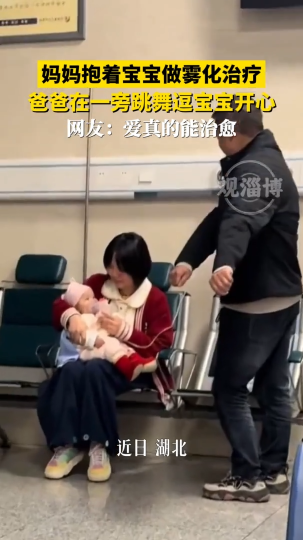 妈妈抱着宝宝做雾化治疗，爸爸在一旁跳舞逗宝宝开心